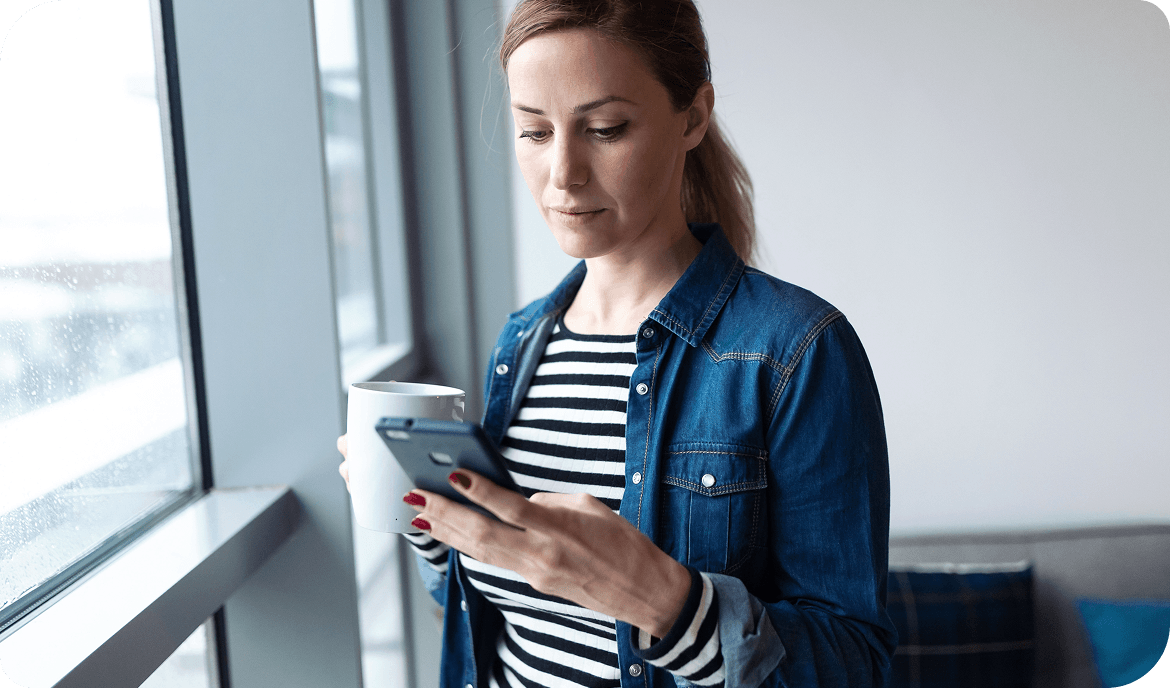 Woman in denim jacket and striped shirt stands by a window with a mug in one hand and looking down at her phone in the other hand