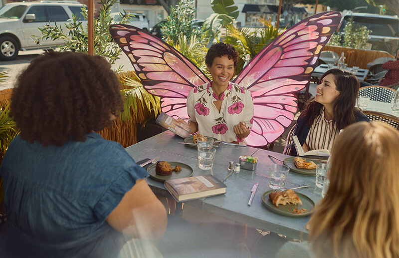 Woman with butterfly wings is sitting at table with other women at a table with food and while holding books
